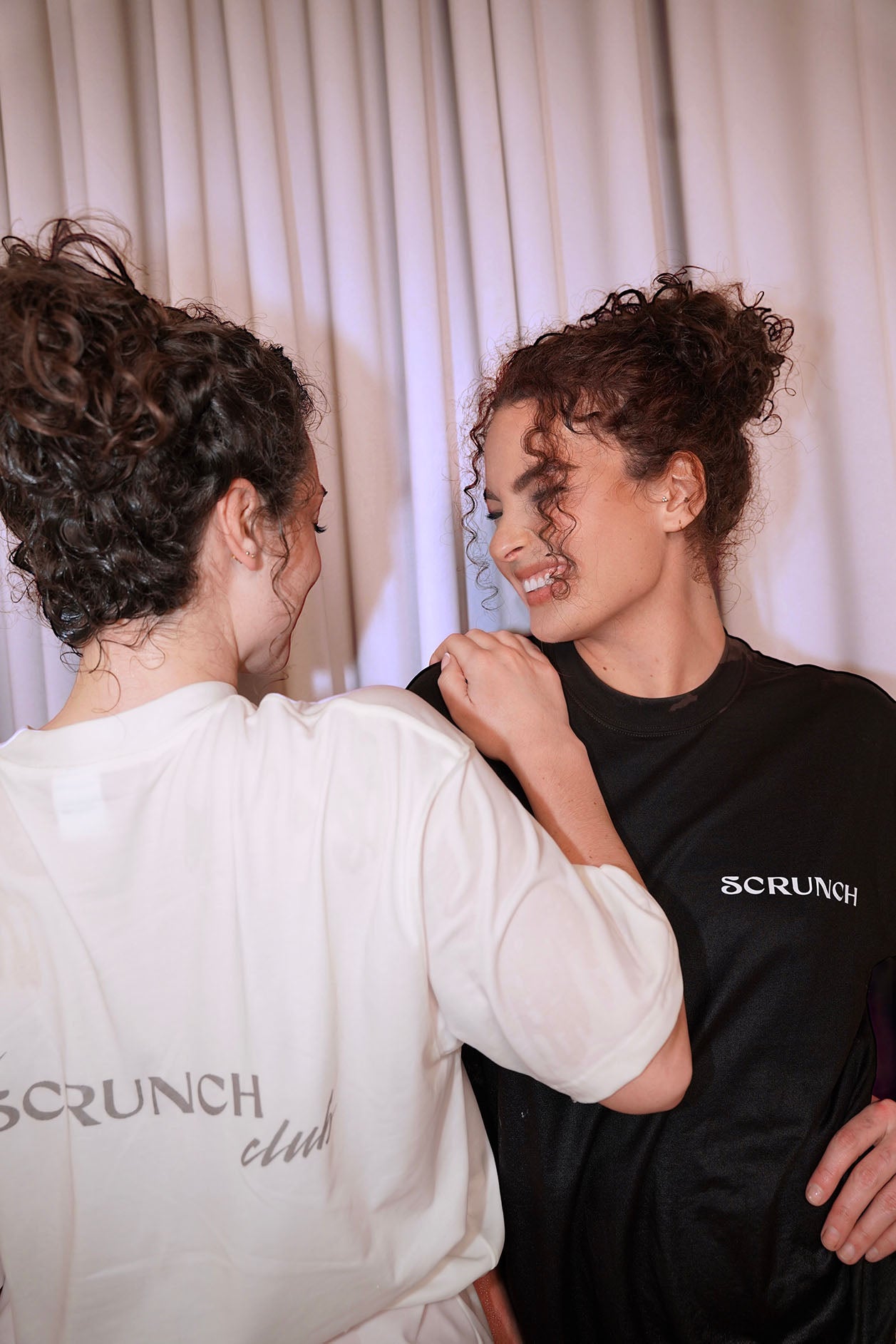 Two people wearing 'Scrunched' t-shirts against a white curtain background