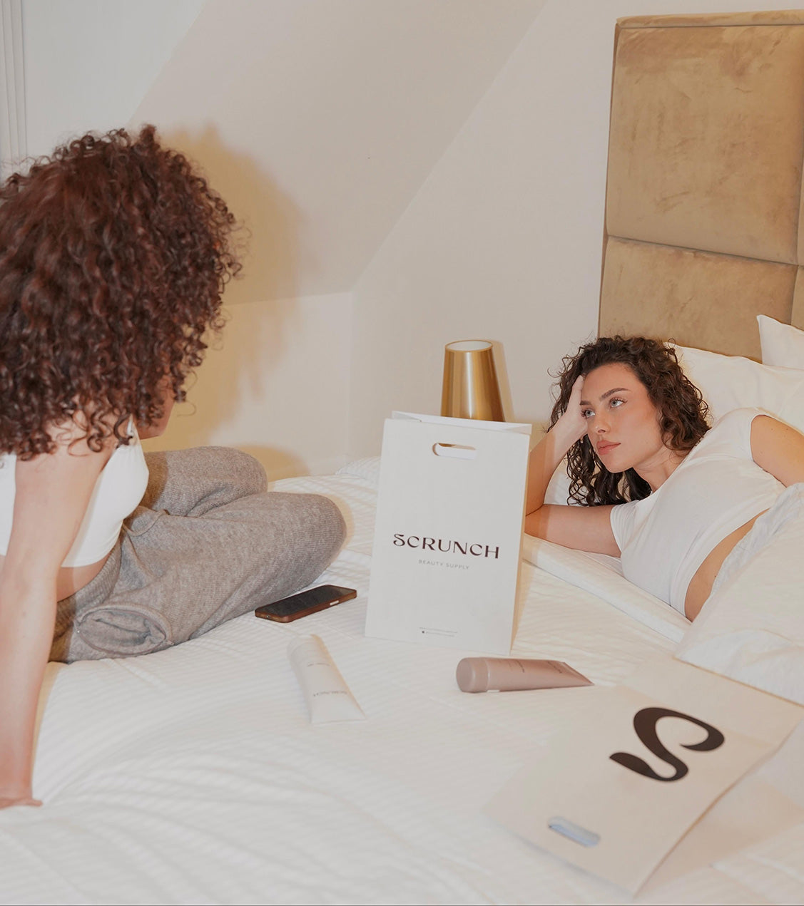 Two women lying on a bed with 'SCRUNCH' branded bags.