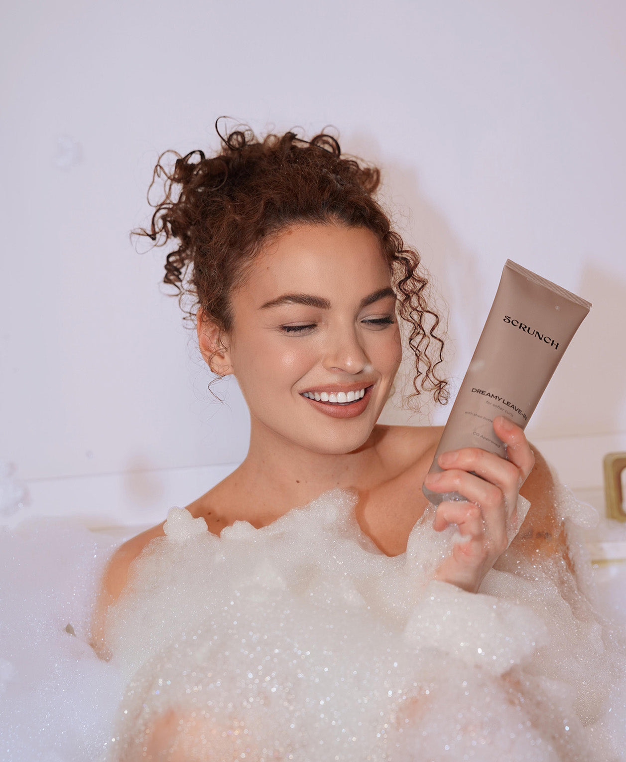 Woman in a bubble bath holding a tube of SCRUNCH CURLY HAIR product.